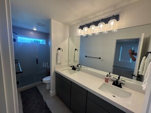 Un baño moderno con un espejo grande, tocador blanco, armarios oscuros y una cabina de ducha de vidrio.