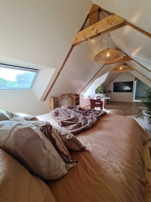 Un dormitorio acogedor en el ático, con techo inclinado, un ventanal grande, una cama con manta y una lámpara colgante.