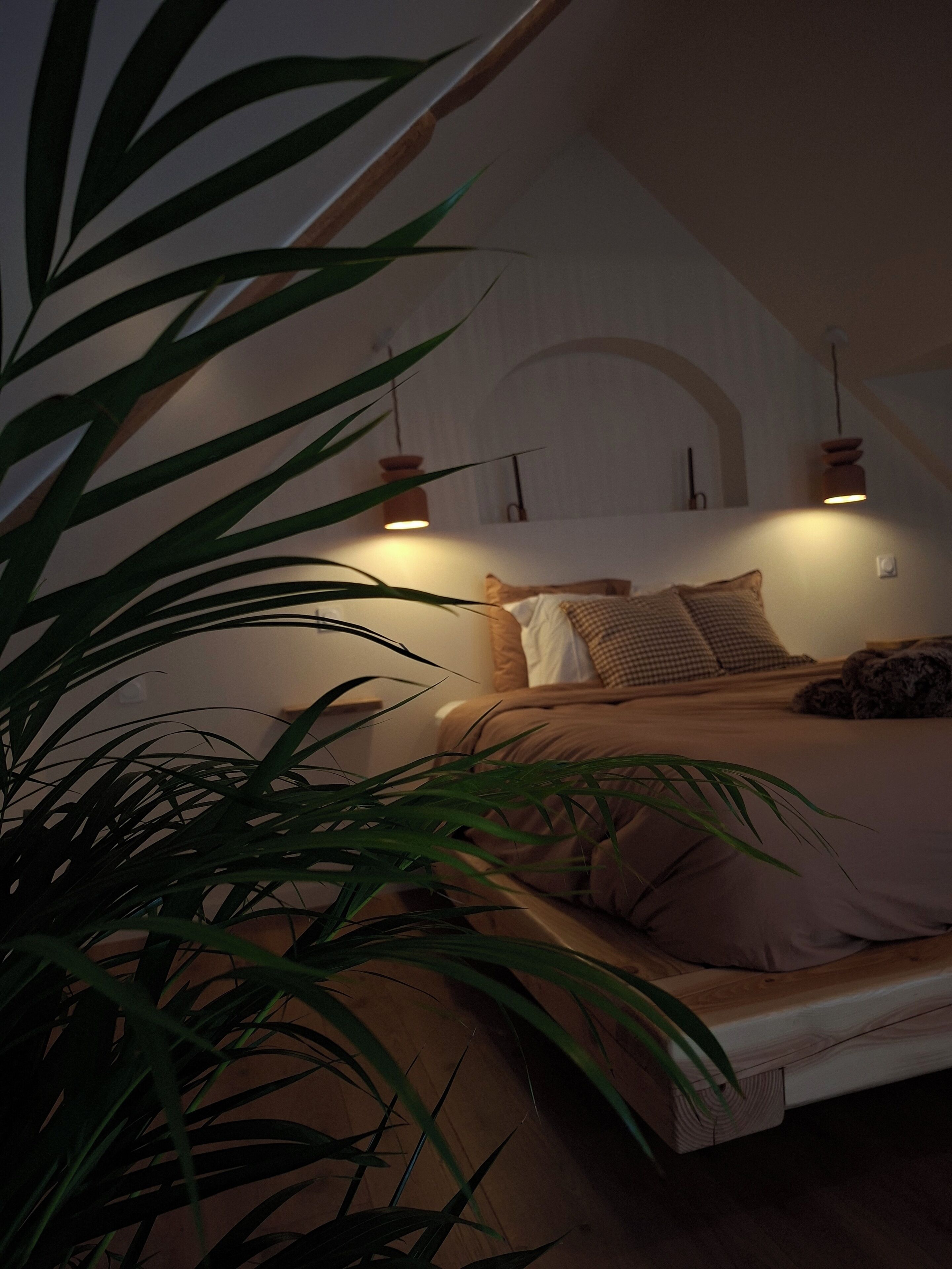 A bedroom with a bed, pillows, a hanging light fixture, and a potted plant.