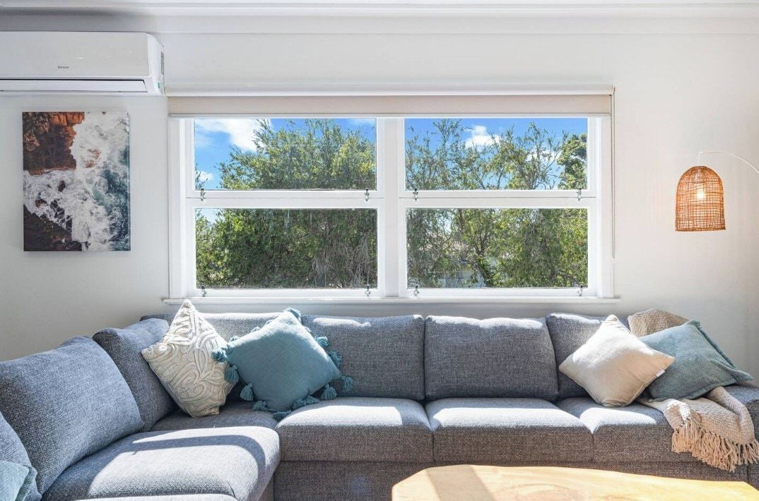 Ein Wohnzimmer mit grauem Sofa, einem Fenster mit Blick auf Bäume und einer Wand-Klimaanlage.