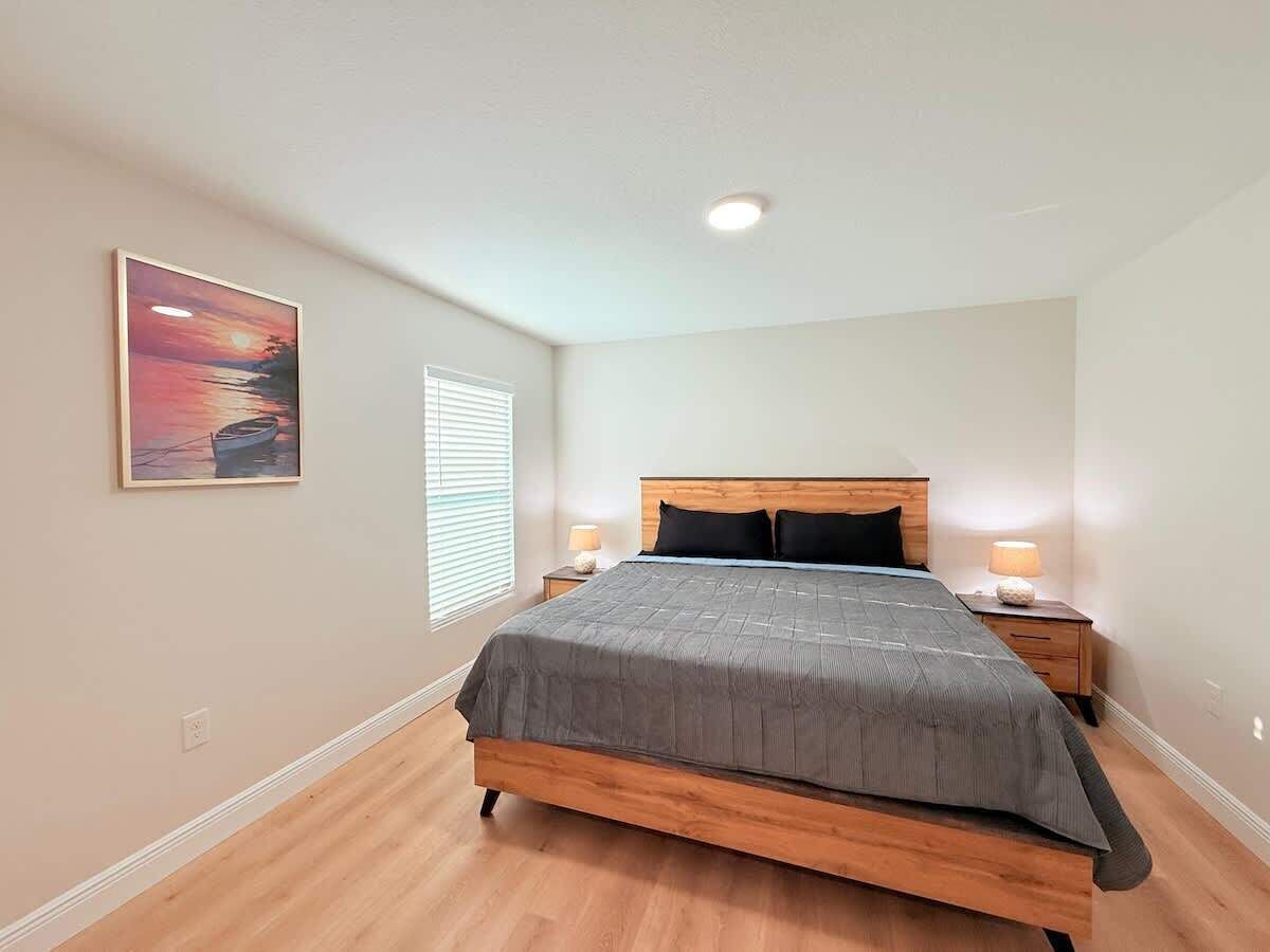 A bedroom with a bed, two bedside tables, a nightstand, a window with blinds, and a painting on the wall.
