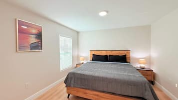 A bedroom with a bed, two bedside tables, a nightstand, a window with blinds, and a painting on the wall.