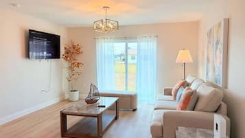A modern living room with a flat-screen TV mounted on the wall, a wooden coffee table, a beige sofa with decorative pillows, a floor lamp, and a large window with curtains.