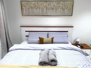 Un lit bien fait, avec une literie à rayures et une serviette pliée, une table de chevet avec une lampe et une télécommande, et un tableau encadré au mur.