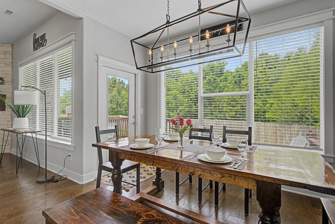 A dining area with a wooden table set for four, a modern chandelier, and large windows with blinds.