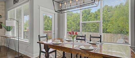Comedor con mesa de madera dispuesta para cuatro, una lámpara moderna y ventanas amplias con persianas.