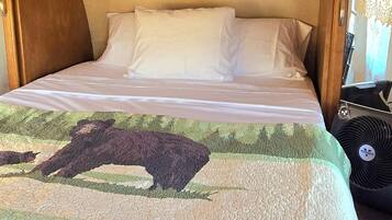 A bed with a quilted cover, a wooden headboard, and a shelf above the bed with books and a small figurine.