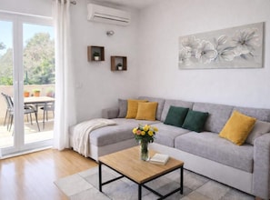 A living room with a grey sofa, yellow and green cushions, a wooden coffee table, and a vase with flowers. There is a balcony with outdoor furniture and plants. - Ljubelle Premium Stay (Seget)