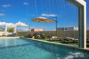 Una piscina con sillas de descanso y sombrillas, una zona de estar cubierta y vistas a montañas al fondo.