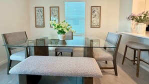 Un comedor con una mesa de vidrio, cuatro sillas, un banco y un jarrón de flores.