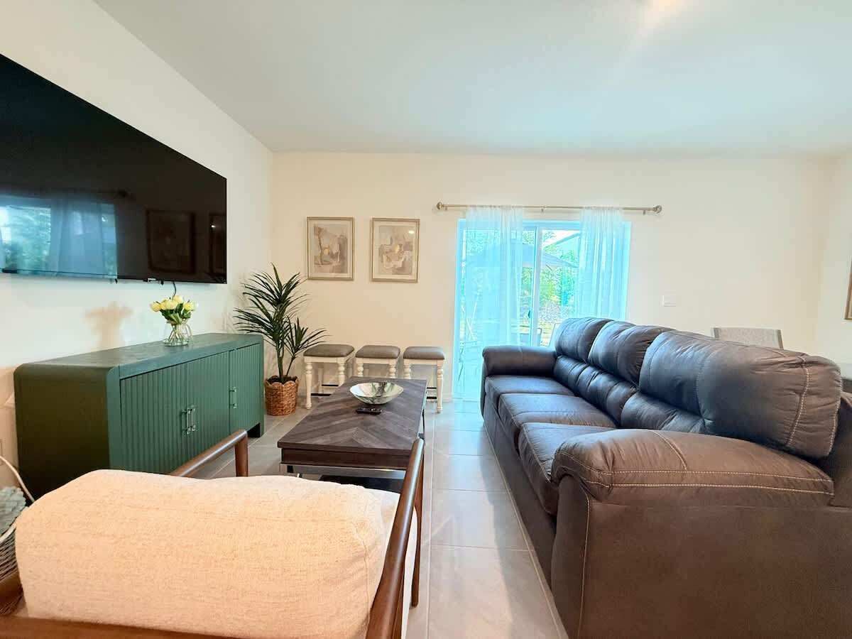 Uma sala de estar com um sofá de couro marrom, um aparador verde, uma mesa de jantar com cadeiras e uma TV fixada na parede.