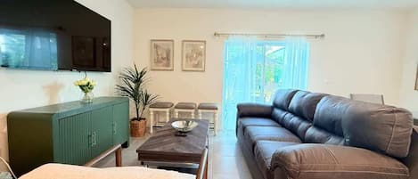 Un salon avec un canapé en cuir marron, un meuble vert, une table à manger avec des chaises et un téléviseur fixé au mur.