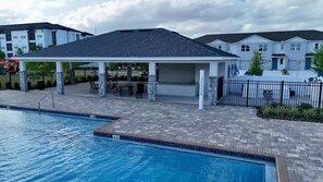 A swimming pool with a covered patio area, a brick walkway, and residential buildings in the background. - Tamarack Magic 3/2 Luxury & Magic Near Disney (Davenport)