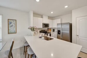 A modern kitchen with white cabinets, stainless steel appliances, a central island, and a dining area with stools. - Tamarack Magic 3/2 Luxury & Magic Near Disney (Davenport)