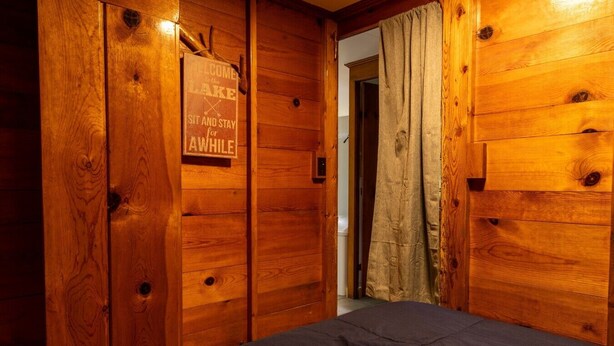 Une pièce en bois avec un lit, une pancarte sur laquelle on peut lire « Bienvenue au bord du lac, installez-vous et restez un peu », et un rideau.