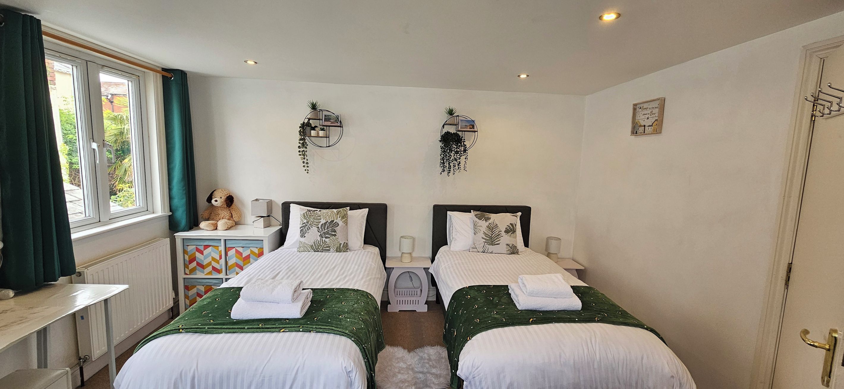 A twin bedroom with two beds, a window with curtains, a small table with a teddy bear, and wall-mounted plants.