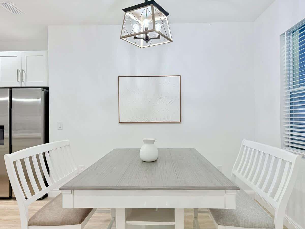 Een eetgedeelte met een rechthoekige tafel, witte stoelen, een witte vaas en een lijst met een afbeelding aan de muur.