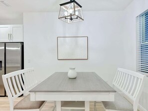 A dining area with a rectangular table, white chairs, a white vase, and a framed picture on the wall.