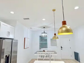 Una cocina moderna con armarios blancos, un refrigerador de acero inoxidable, una mesa de comedor con una silla blanca e iluminación colgante.