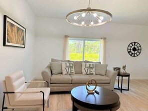 Sala de estar com sofá, poltrona e mesa de centro. Há uma janela com cortinas, um relógio de parede e um quadro emoldurado.