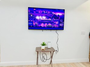 Uma televisão de tela plana presa a uma parede branca, uma pequena mesa de madeira com uma planta em vaso e um alto-falante branco, e um cabo de alimentação.
