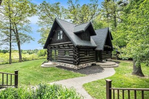 A log cabin with a steeply pitched roof, surrounded by a well-maintained lawn and trees.