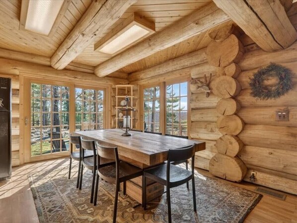 A wooden cabin interior with a dining table, chairs, a fireplace, and a large window.