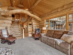 A log cabin interior with a staircase, leather furniture, and large windows.