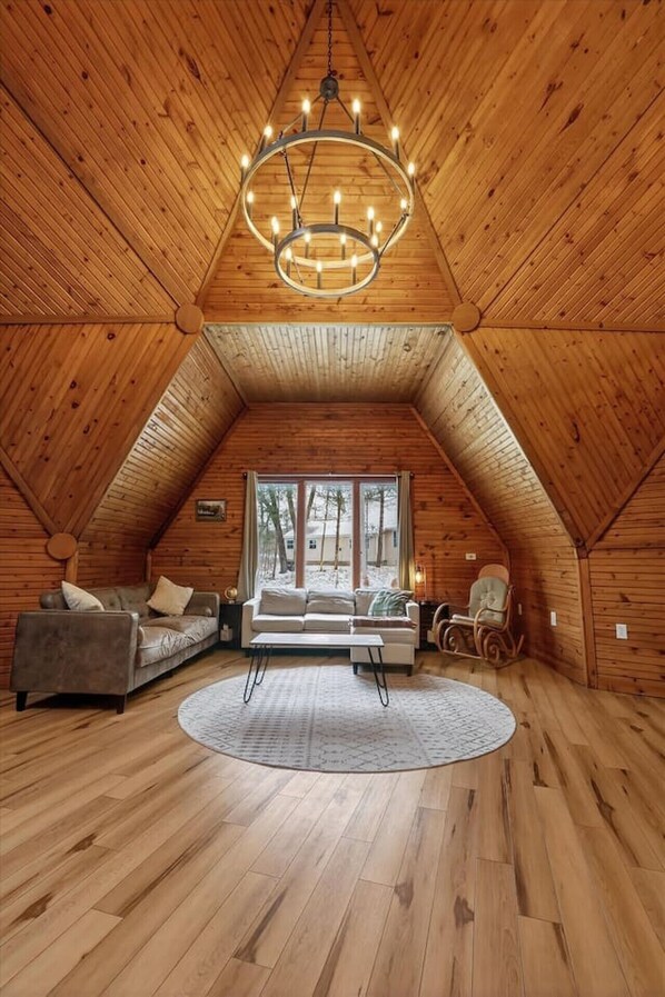 A cozy attic room with wooden walls, a large window, a sofa, a rocking chair, and a chandelier.