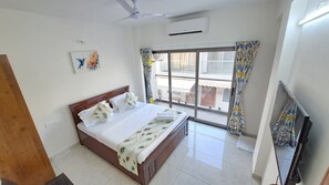 A hotel room with a bed, a television, a ceiling fan, a window with curtains, and an air conditioning unit.