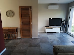Une pièce avec une porte en bois, un téléviseur posé sur un meuble blanc et une grande fenêtre.