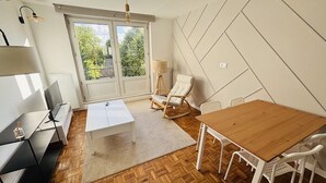 Un soggiorno moderno con pavimento in legno, un divano bianco, un tavolo da pranzo con sedie bianche, una poltrona a dondolo e un'ampia finestra vista verde.