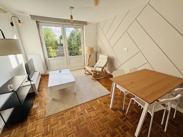 Un salon moderne avec un plancher en bois, un canapé blanc, une table à manger avec des chaises blanches, un fauteuil à bascule et une grande fenêtre donnant sur un paysage verdoyant.