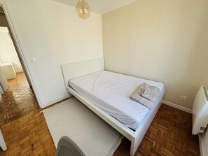 Una habitación pequeña y limpia con una cama individual, una silla, un radiador y una lámpara colgante.