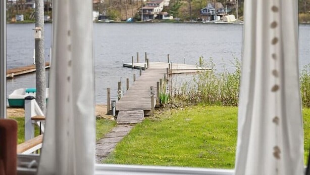 Et værelse med et stort vindue der kigger ud over en sø, en træbro og en græsplæne.