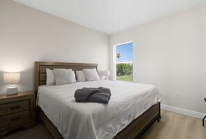 A bedroom with a wooden bed, white bedding, a nightstand with a lamp, and a window with blinds. - Chic 3BR, 2BA – Brand New! (Englewood)