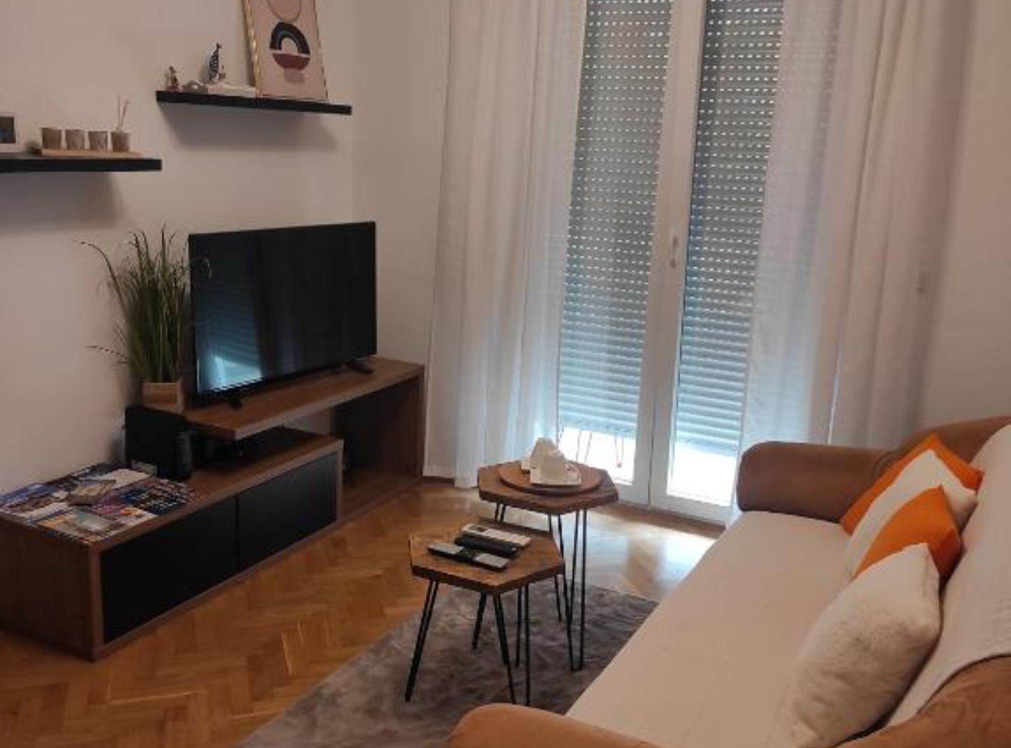 Una sala de estar con una televisión de pantalla plana, piso de madera, un sofá, una mesita auxiliar y una ventana con persianas.
