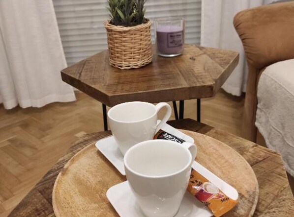 Et træbord med to hvide kopper og en kaffepakke, en fletkurv med en plante, og et lys på et lille træbord.