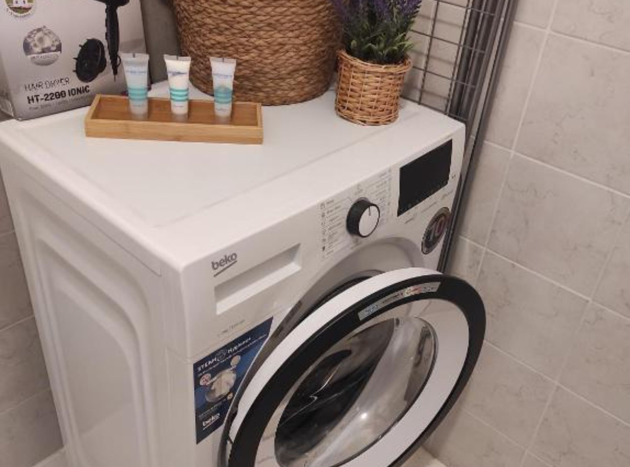 A front-loading washing machine with a control panel, a wooden tray with hair care products, and a woven basket with a plant.