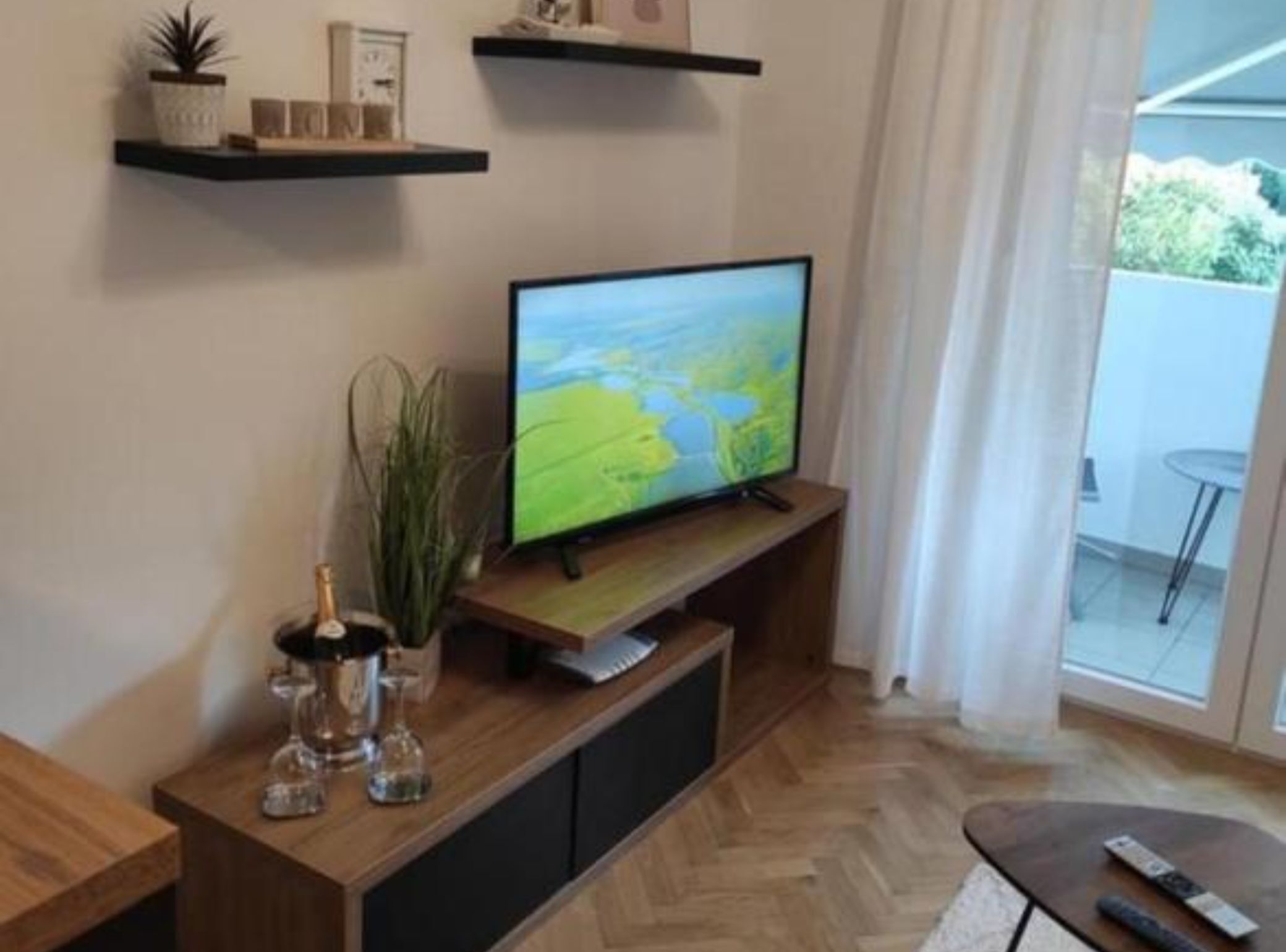 Een woonkamer met een flatscreen televisie op een houten standaard, een bijzettafeltje met een afstandsbediening, en een potplant.