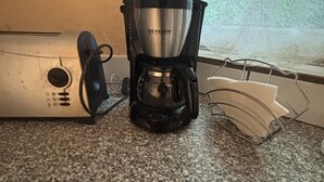 A coffee maker on a granite countertop near a window with lace curtains.