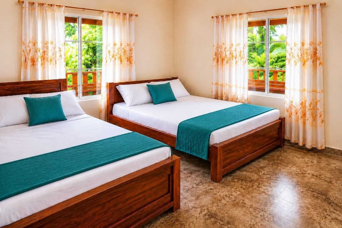 Two wooden beds with white linens and teal accents, large windows with floral curtains, and a tiled floor.