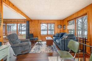 A cozy cabin interior with wooden walls, a dining area, a sofa, and a view of a lake.