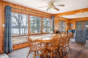 A wooden interior with a dining table, chairs, a sofa, and large windows.