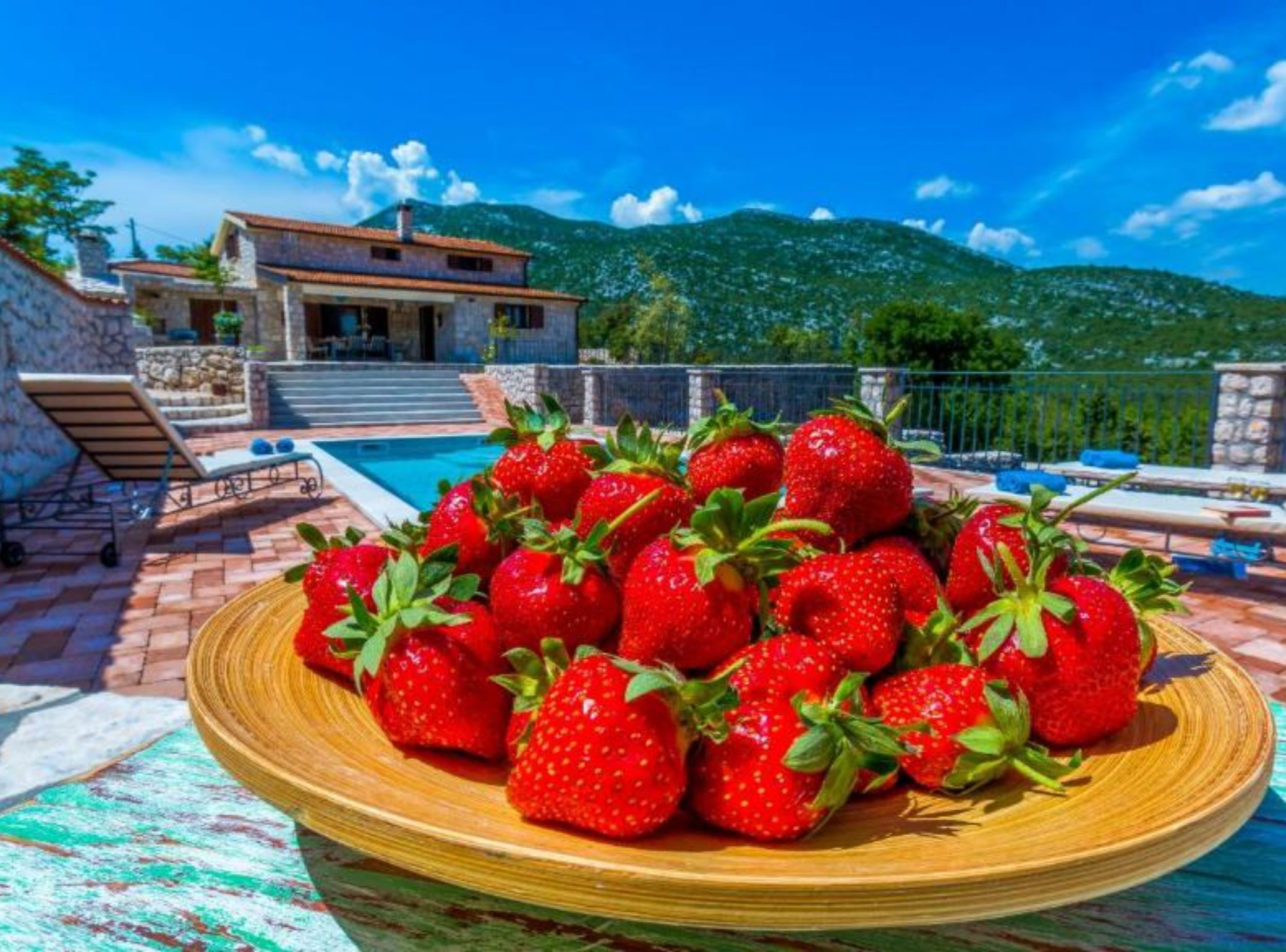 A poolside setting with a plate of strawberries, a stone house, and a mountainous backdrop.