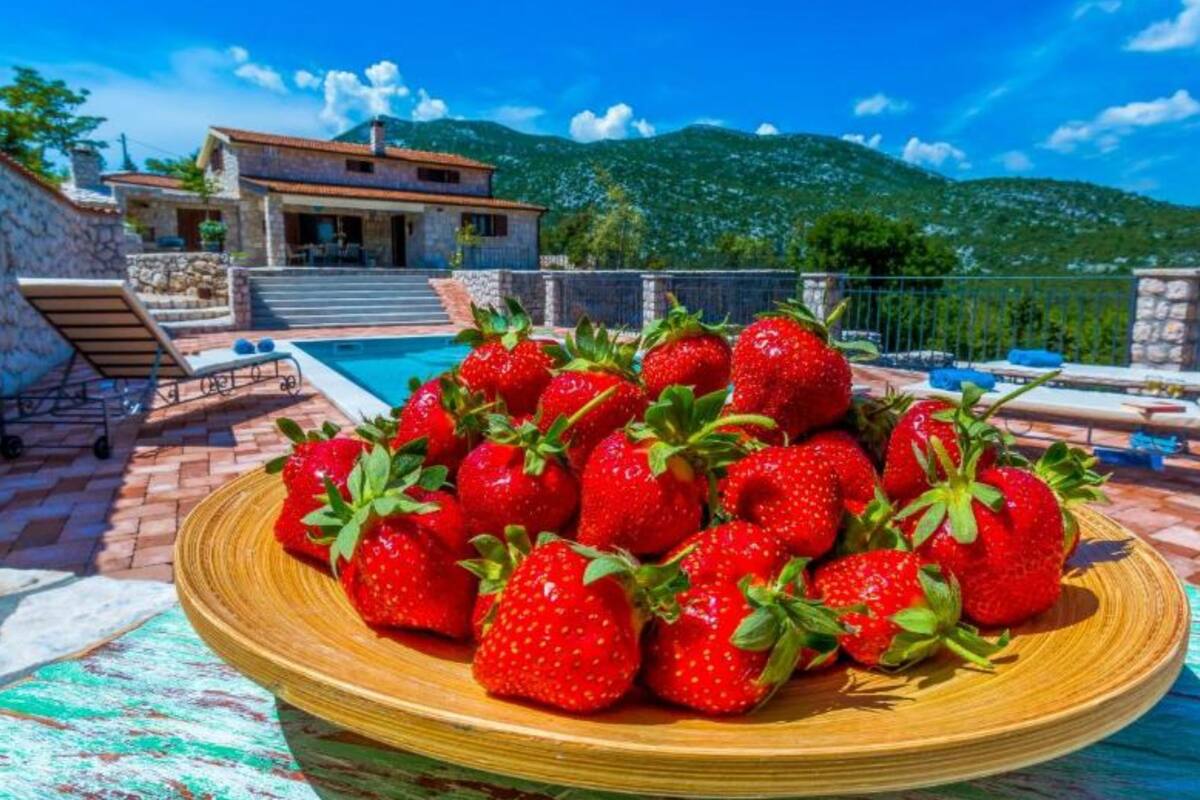A poolside setting with a plate of strawberries, a stone house, and a mountainous backdrop.