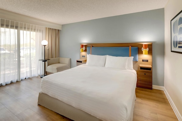 Une chambre d’hôtel avec un grand lit, une chaise, une table de chevet avec une lampe et une fenêtre avec des rideaux.