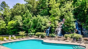 A rectangular swimming pool with a waterfall feature, surrounded by lounge chairs and a wooded area. - Sunset Views • Hot Tub • Pool Table • Sleeps 5 (Sevierville)