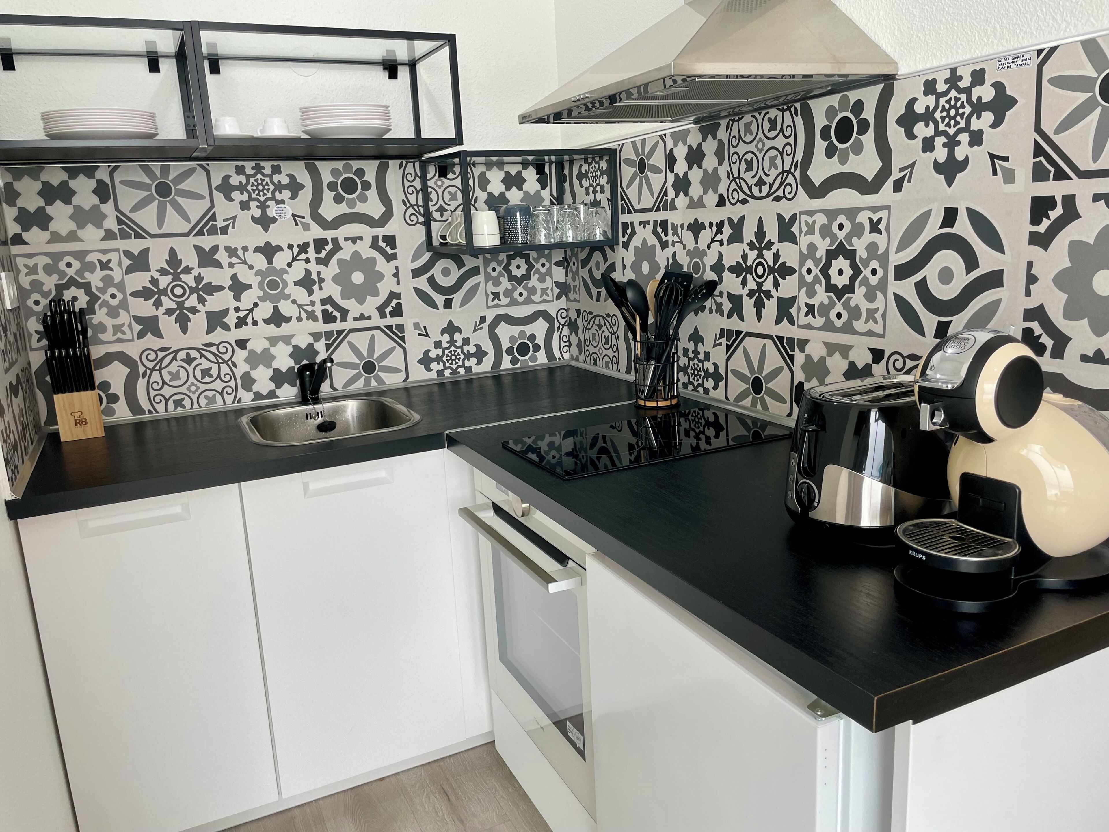 A kitchen with a black countertop, white cabinets, a stainless steel sink, a coffee maker, and patterned backsplash tiles.