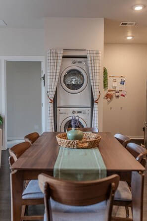 A dining area with a table, chairs, a central vase, and a washing machine in the background. - Sipped Away: King Beds, Walk in Closets (Traverse City)